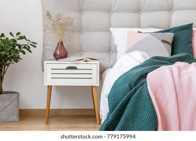 Close-up Of Dark Vase With Flowers On White Nightstand Next To Bed With Pink And Green Bedsheets In Pastel Bedroom Interior