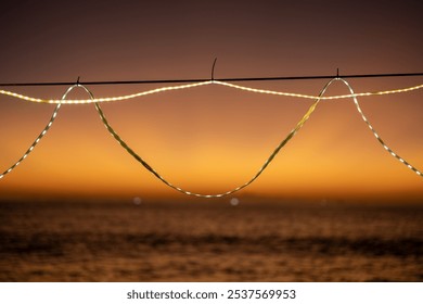 Close-up of decorative string lights hanging against a warm, glowing sunset over the ocean. The lights create a soft and cozy ambiance, contrasting with the fading daylight. - Powered by Shutterstock