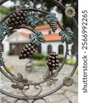 A close-up of decorative wrought iron artwork featuring grape clusters and vine leaves with intricate details, set against the blurred background of a traditional building and courtyard. The artistic 