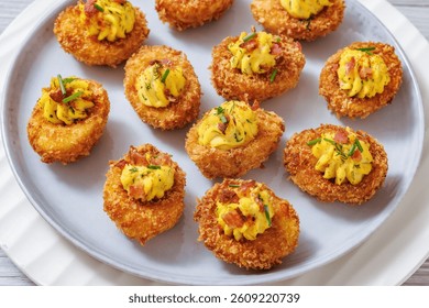 close-up of Deep fried deviled eggs with crispy coating of panko and parmesan, sprinkled with fresh chives and crumbled bacon on white plate on wooden table