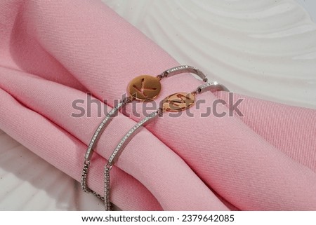 A close-up of delicate bracelets with gold circular pendants engraved with the letters 'K' and 'M', gracefully laid upon soft pink fabric with a rippled white background
