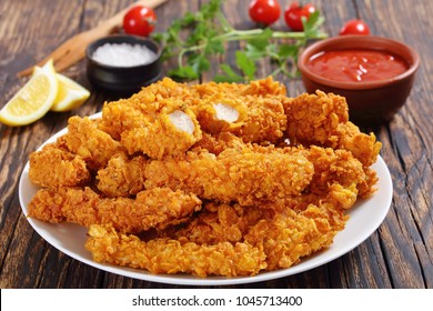 close-up of delicious crispy fried chicken breast strips on white plate, on old rustic  wooden table with tomato sauce and lemon slices, easy recipe for outdoor picnic or party, view from above