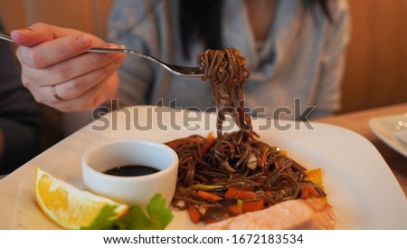 Close-up of delicious dish of buckwheat noodles with salmon and soy sauce on white plate. Woman's hand twisting noodles on fork.
