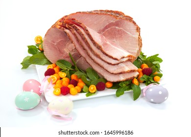 Closeup of delicious roasted sliced Easter ham with fresh mint, kumquat, and raspberries over white background. Easter eggs.