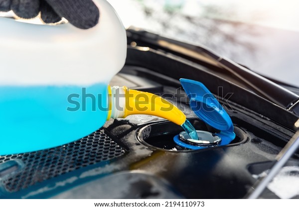 Close-up detail of driver or mechanic hand in
gloves pouring blue antifreeze liquid for winter car windshield
screen washing with bootle watering can. Car seasonal service and
maintenance concept