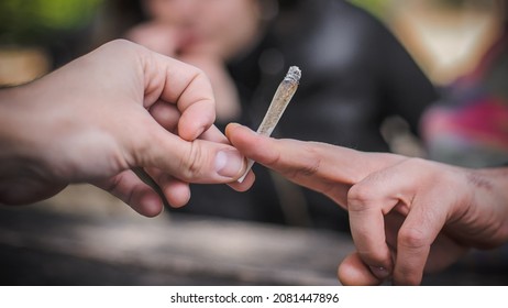 Closeup Detail Macro View Of Marijuana Joint Circling Around From Hand To Hand. Young Friends Smoking Cannabis Marijuana Ganja Or Hashish Cigarette. Real Life Scene Concept