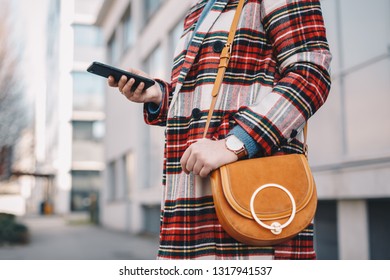 Close-up Detail Of Modern Day Female Accessories - Stylish Young Woman Wearing An Overcoat With A Tartan Pattern And A Wrist Watch While Holding A Fancy Camel Bag And Typing On Her Mobile Phone.