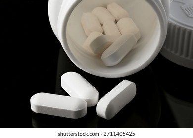 Close-up of detailed white oval or rectangular pills stacked in the mouth of a white container, with some scattered on a dark reflective background.