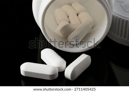 Close-up of detailed white oval or rectangular pills stacked in the mouth of a white container, with some scattered on a dark reflective background.