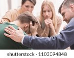 Close-up of a devastated young man holding his head in his hands and friends supporting him during group therapy