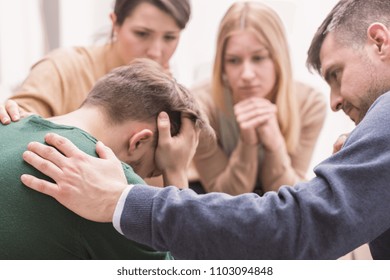 Close-up of a devastated young man holding his head in his hands and friends supporting him during group therapy - Shutterstock ID 1103094848