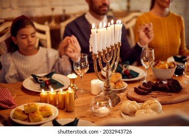 Close-up of dining table with lighted candles in menorah at dining table on Hanukkah. Family in the background is holding hands and praying. - Powered by Shutterstock