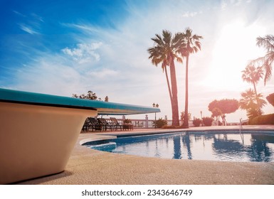 Close-up of a diving board over the open swimming pool during sunset light through south palm trees on hot summer evening