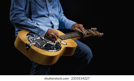 Close-up of a Dobro guitar resting on the lap of a musician dressed in cowboy attire on a dark backdrop - Powered by Shutterstock