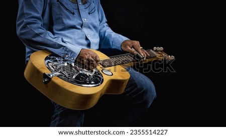 Close-up of a Dobro guitar resting on the lap of a musician dressed in cowboy attire on a dark backdrop