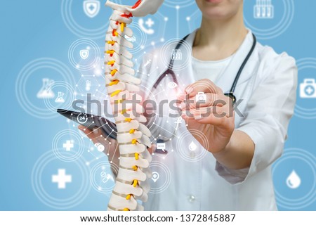 A closeup of doctor with stethoscope operating with artificial spine model holding a tablet in hand inside a medical service structure. The concept of innovative approach in spine diseases treatment.