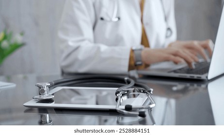 Close-up of doctor's desk with stethoscope and tablet computer. Physician is using a laptop at the background. Medicine concept - Powered by Shutterstock