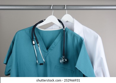 Closeup of a doctor's scrubs with stethoscope and lab coat on hangers against a neutral background. Green Scrubs and a white lab coat against a gray background.