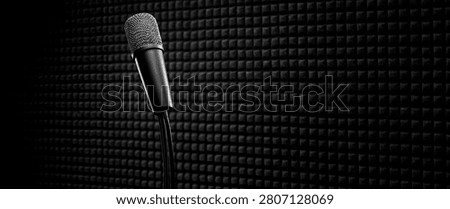 A close-up, dramatic black and white shot of a professional microphone positioned on a stand against a textured, sound-dampening wall.