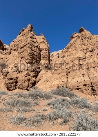 Close-up of dramatic red and orange eroded rock spires rising against a deep blue sky in Fairy Tale Canyon (Skazka), Kyrgyzstan. The heavily textured sandstone formations with sharp pointed peaks resemble ancient ruins or fantasy castle towers. Sparse dry desert shrubs at the base.