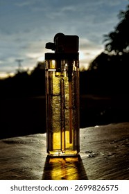 A close-up, dramatic shot of a clear, disposable plastic lighter standing upright on a rough, wooden table or surface. The lighter is strongly backlit by the setting sun during the golden hour.