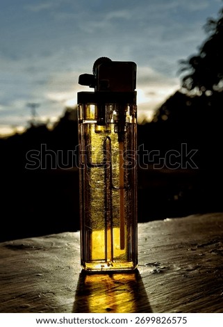 A close-up, dramatic shot of a clear, disposable plastic lighter standing upright on a rough, wooden table or surface. The lighter is strongly backlit by the setting sun during the golden hour.