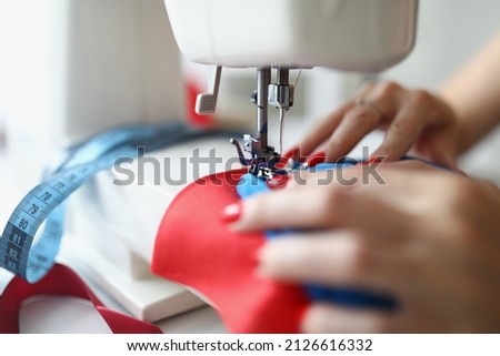 Close-up of dressmaker lady working on white sewing machine. Professional seamstress creating red trendy dress. Process of tailor at work. Workshop and fashion factory concept