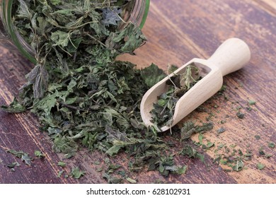 Closeup Of Dried Nettle Leaves In Glass With Wooden Spoon On Scratched, Dark Wood