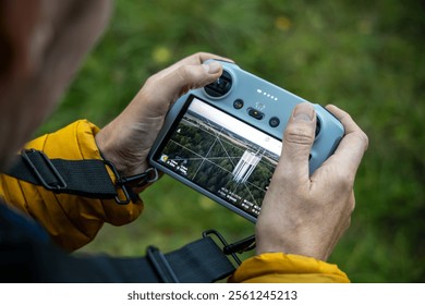 Close-up of a drone remote controller held in hands, showing the screen with a live view of a water tower and surrounding forest from the drone camera during flight.