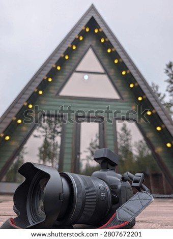 Closeup DSLR camera with wideangle lens on wooden deck, set before triangular cabin facade with warm string lights, green siding, reflective windows, and soft dusk light in outdoor retreat setting