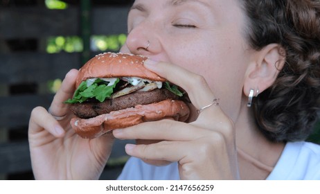 Close-up Eating Fast Food. A White Woman Takes A Big Bite From An Appetizing Burger And Enjoys Chewing Delicious Food, Getting Dirty With Sauce. An Unhealthy Diet Leads To Heart Disease.