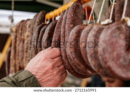 Close-up of an elderly hand touching rows of traditional cured sausages hanging on display at an outdoor farmers market. The image conveys authenticity, craftsmanship, and connection to local food traditions and artisanal meat production.