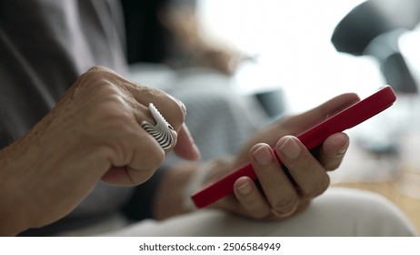 Close-up of elderly woman’s hands using a red smartphone, wearing a ring, demonstrating senior interaction with modern technology, focus on hands, digital communication, indoor home setting