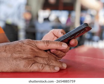 Close-up of elderly hands using a smartphone on red table at an outdoor restaurant, with soft blurry background. Concept of senior tech use, waiting moment, and modern digital lifestyle.
