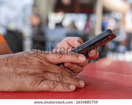 Close-up of elderly hands using a smartphone on red table at an outdoor restaurant, with soft blurry background. Concept of senior tech use, waiting moment, and modern digital lifestyle.
