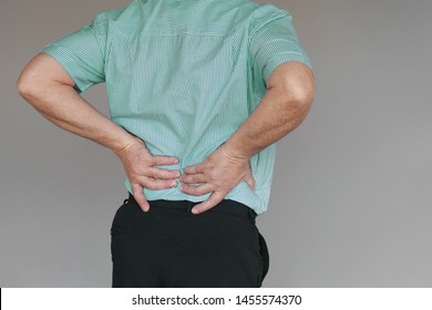 Close-up Elderly Man With Pain In Kidneys Isolated On A Gray Background. Old Man With Back Ache Clasping Her Hand To Her Lower Back. Man Suffering From Ribbing Pain, Waist Pain.