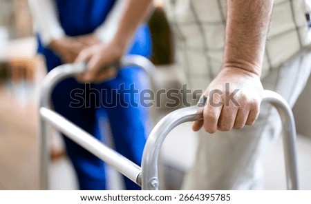 Close-up of elderly man using a walker with caregiver in blue scrubs providing support at home.