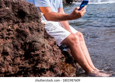 Close-up Of An Elderly Man's Bare Legs Using Smart Phone Sitting On The Rocks Enjoying The Vacation In The Sea - Social And Technological Senior People Concept