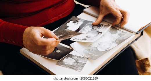 Close-up Of Elderly Woman Holding Old Black And White Film Photo Of Her Childhood While Sitting At Home Alone. Senior Woman Looking At Vintage Photo Album. Selective Focus On Retro Photograph
