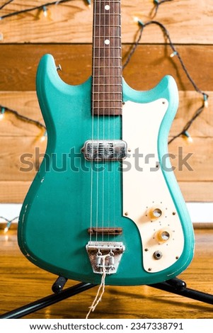 A close-up of an electric teal guitar with worn edges, rustic electric knobs, metal bridge and fretboard on black guitar stand with twinkle lights on wooden wall in the background.