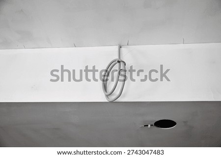 Close-up of the electrical installation stage in an interior renovation. A coil of grey protective corrugated conduit with an electrical cable is attached to the white wall, ready for connection. In the dark grey drywall ceiling in the foreground, a neat circular cutout with protruding wires for a recessed spotlight is visible. The photo illustrates the process of renovation, rough-in finishing, and utility routing