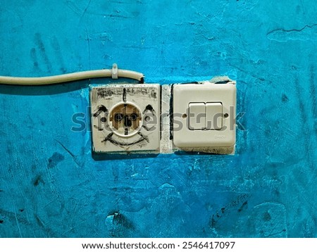 A close-up of an electrical outlet and a light switch mounted on a textured blue wall. The outlet has been decorated with a simple drawn face, giving it a playful and whimsical appearance.