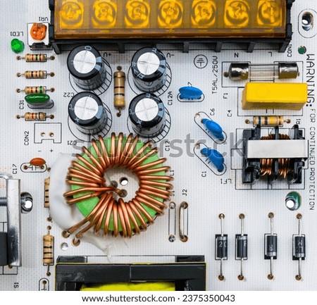 Close-up of electronic components on white printed circuit board. Toroidal coil and various capacitors, electrical fuse and resistor trimmer or diodes on power supply inverter PCB with screw terminal.