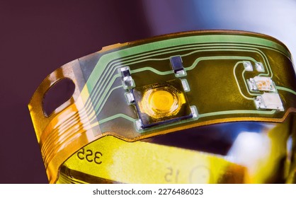 Closeup of electronic flexible printed circuit on plastic strip curled to ring on purple background. Green and yellow lines and small electric components on flex PCB strip from disassembled earphones. - Shutterstock ID 2276486023