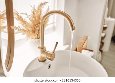 Close-up of an elegant golden faucet in the bathroom sink next to stylish decorations. A beautiful sink with a golden faucet next to an oval mirror and a shelf with hand towels. 