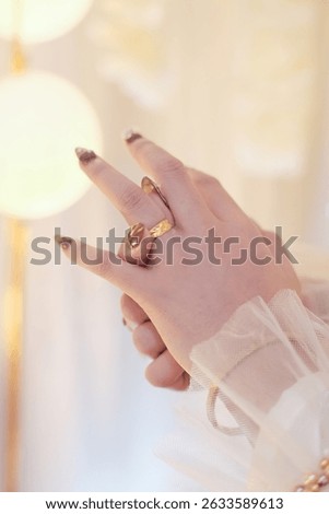 Close-up of elegant hands featuring chunky gold rings and stylish gold and dark tip manicure. Soft, glowing background and sheer fabric cuff add a touch of luxury and femininity. 