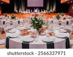 Close-up of an elegant round table setup with a floral centerpiece, plates, glasses, and food dishes in a banquet hall with a stage.