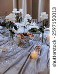 Close-up of an elegant round table setup with a floral centerpiece, plates, glasses, and food dishes in a banquet hall 