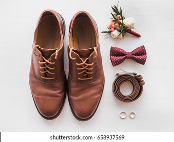 Closeup of elegant stylish brown male accessories isolated on white wooden background. Top view of bow-tie, belt, shoes, floral corsage, golden rings. Preparation for wedding concept. Horizontal photo