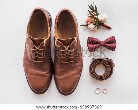 Closeup of elegant stylish brown male accessories isolated on white wooden background. Top view of bow-tie, belt, shoes, floral corsage, golden rings. Preparation for wedding concept. Horizontal photo